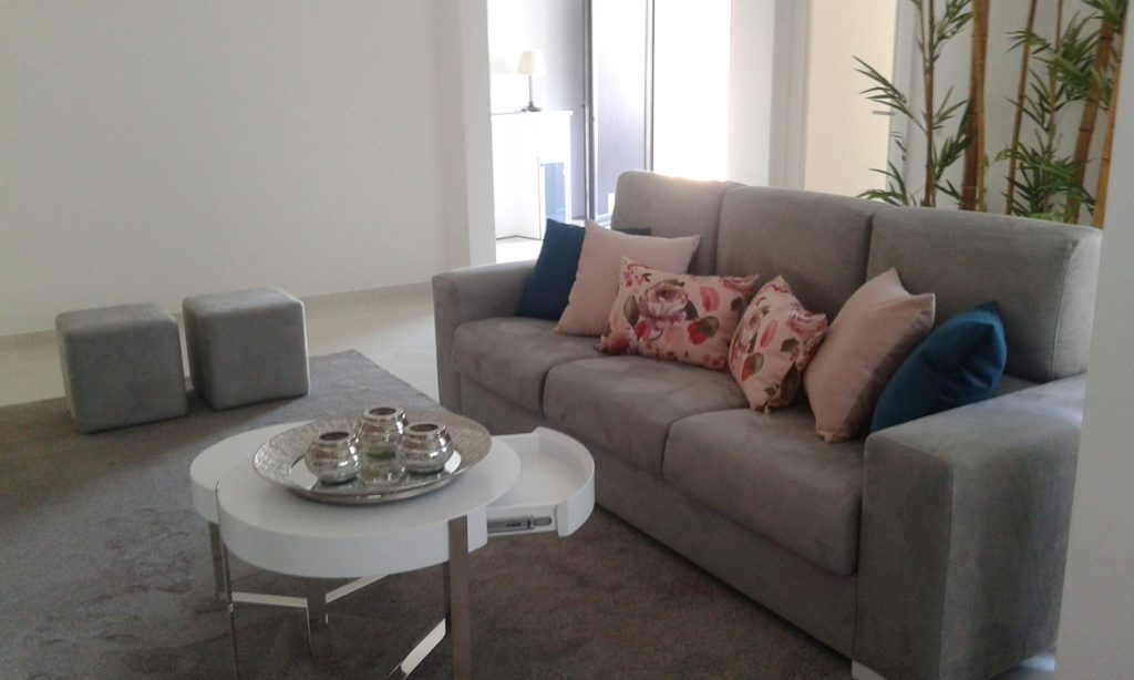 Sala luminosa com sofá cinzento, almofadas rosa e azuis e mesa de centro redonda