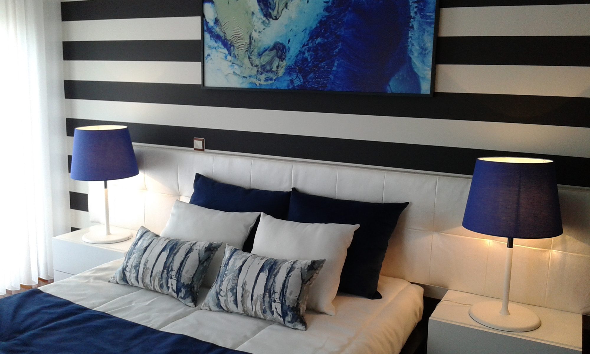 Quarto com parede às riscas pretas e brancas, quadro azul e roupa de cama azul-marinho