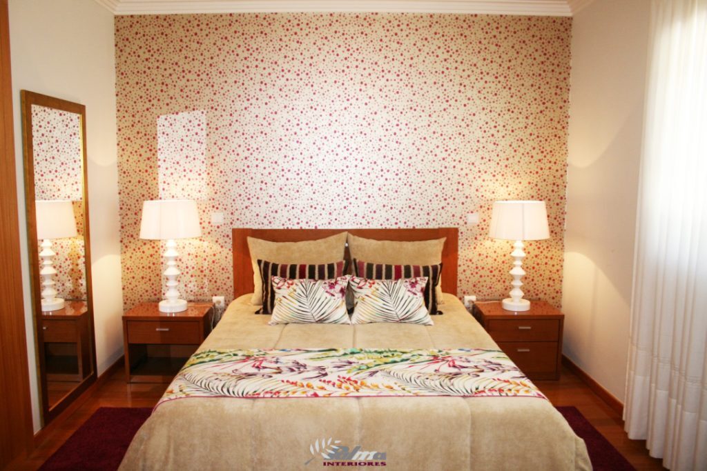 Quarto com parede revestida a papel de parede floral, cabeceira em madeira e candeeiros brancos