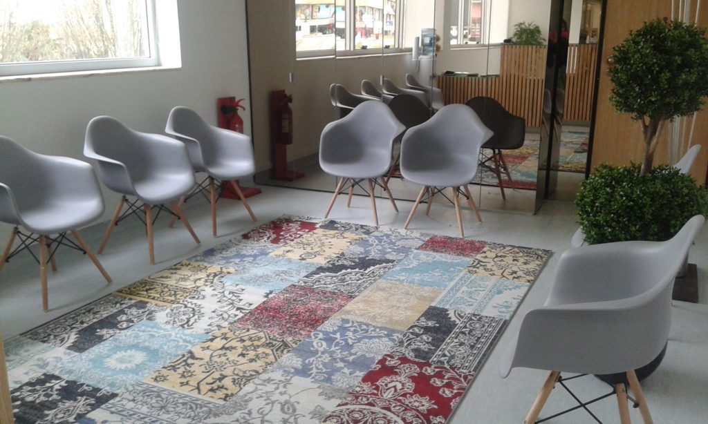 Sala de espera com cadeiras brancas, grandes janelas e tapete patchwork colorido