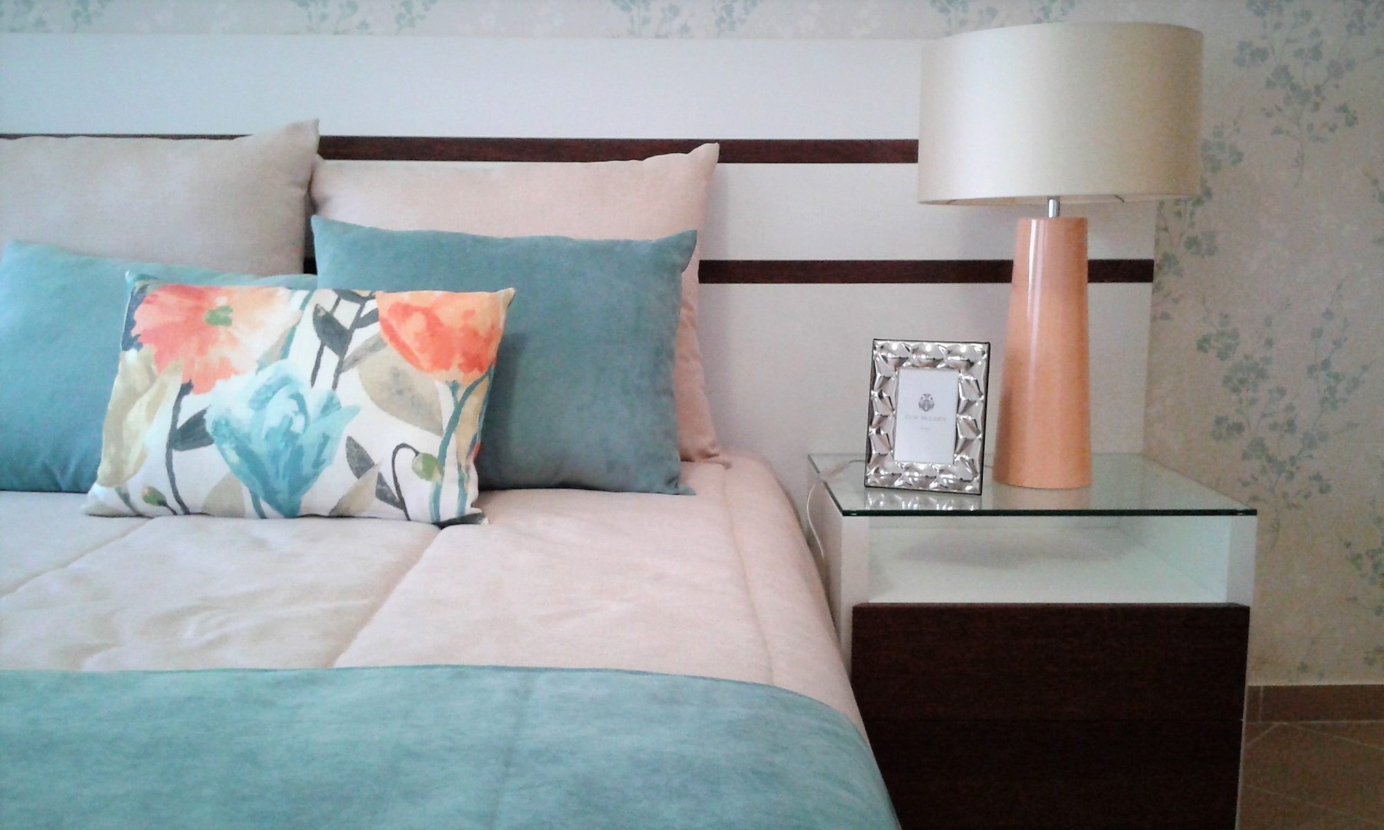 Pormenor de cama com almofadas estampadas, colcha em tons de azul e rosa e candeeiro de mesa